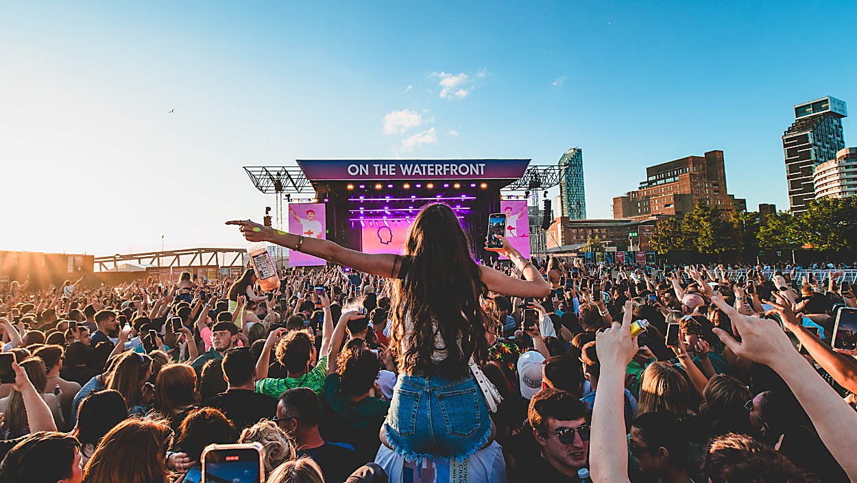 Crowd with hands raised in front of an outdoor waterfront festival stage at sunset